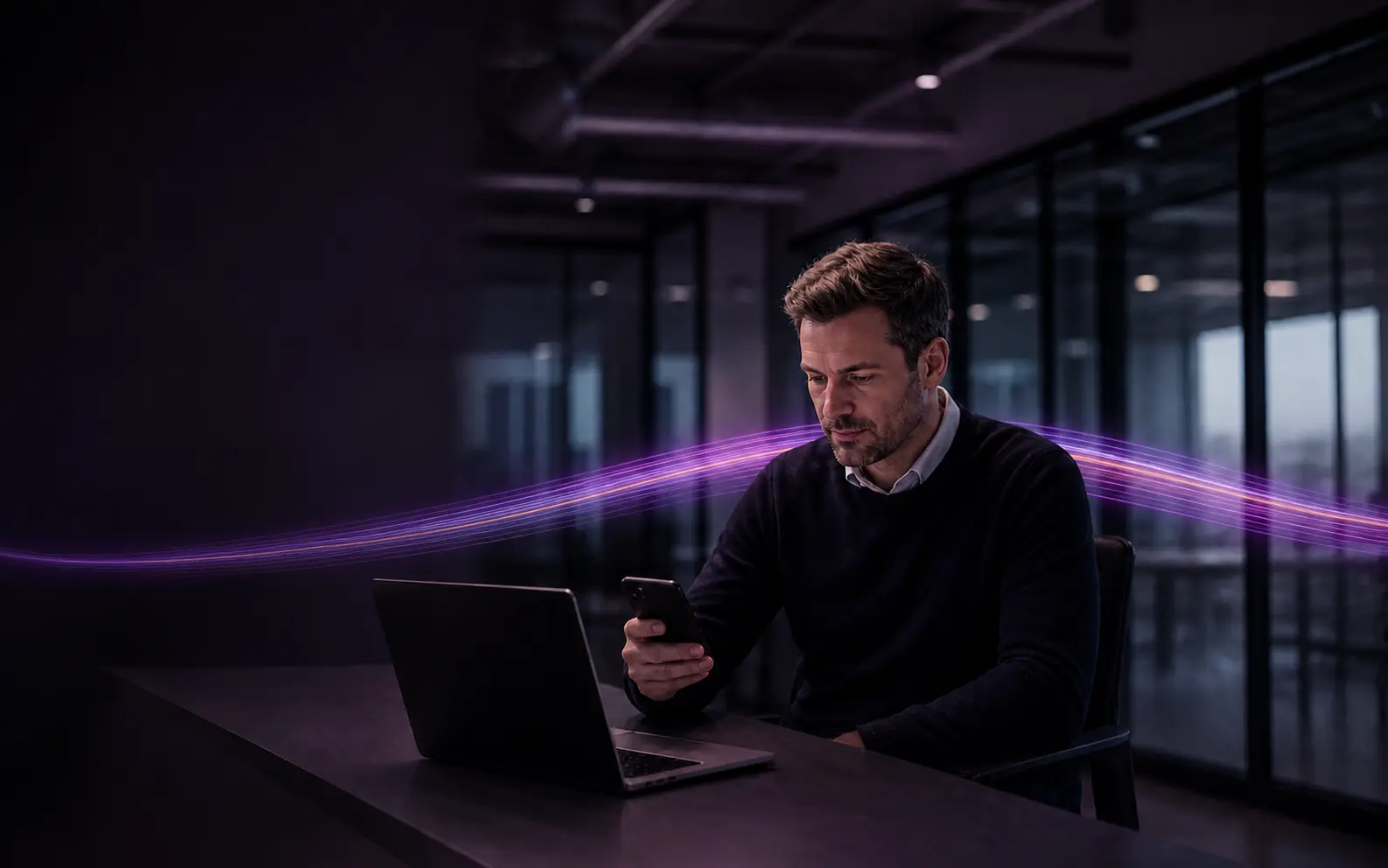 A man at a desk in a darkened modern office checking his mobile, laptop closed beside him, with a glowing violet and magenta ribbon passing along the desk around him.