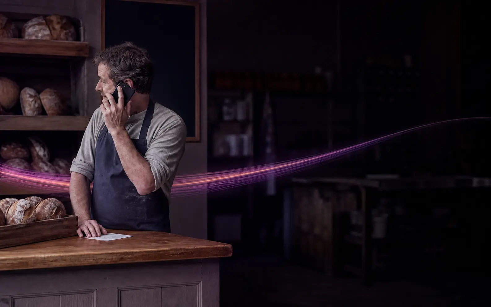 A shopkeeper standing at the counter of a bakery taking a call on his mobile, with a glowing violet and magenta ribbon passing through the shop around him.